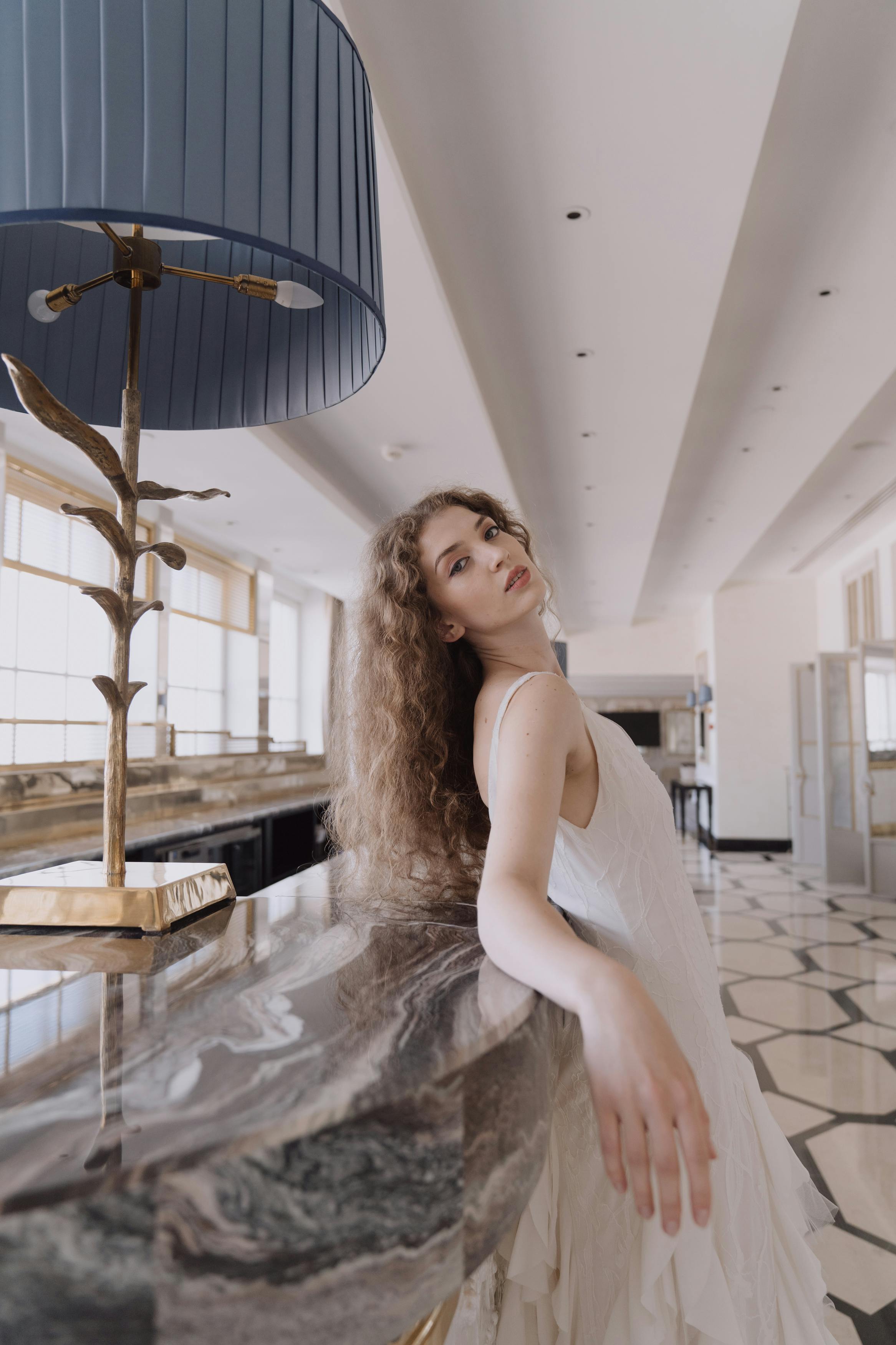 Beautiful woman in a white dress leaning on a marble counter indoors, beside a stylish lamp.