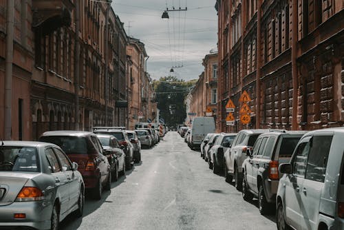 Rows of cars parked near paved road under tall buildings