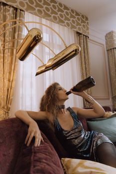Elegant woman in a black dress enjoys wine on a luxurious sofa indoors, exuding sophistication.