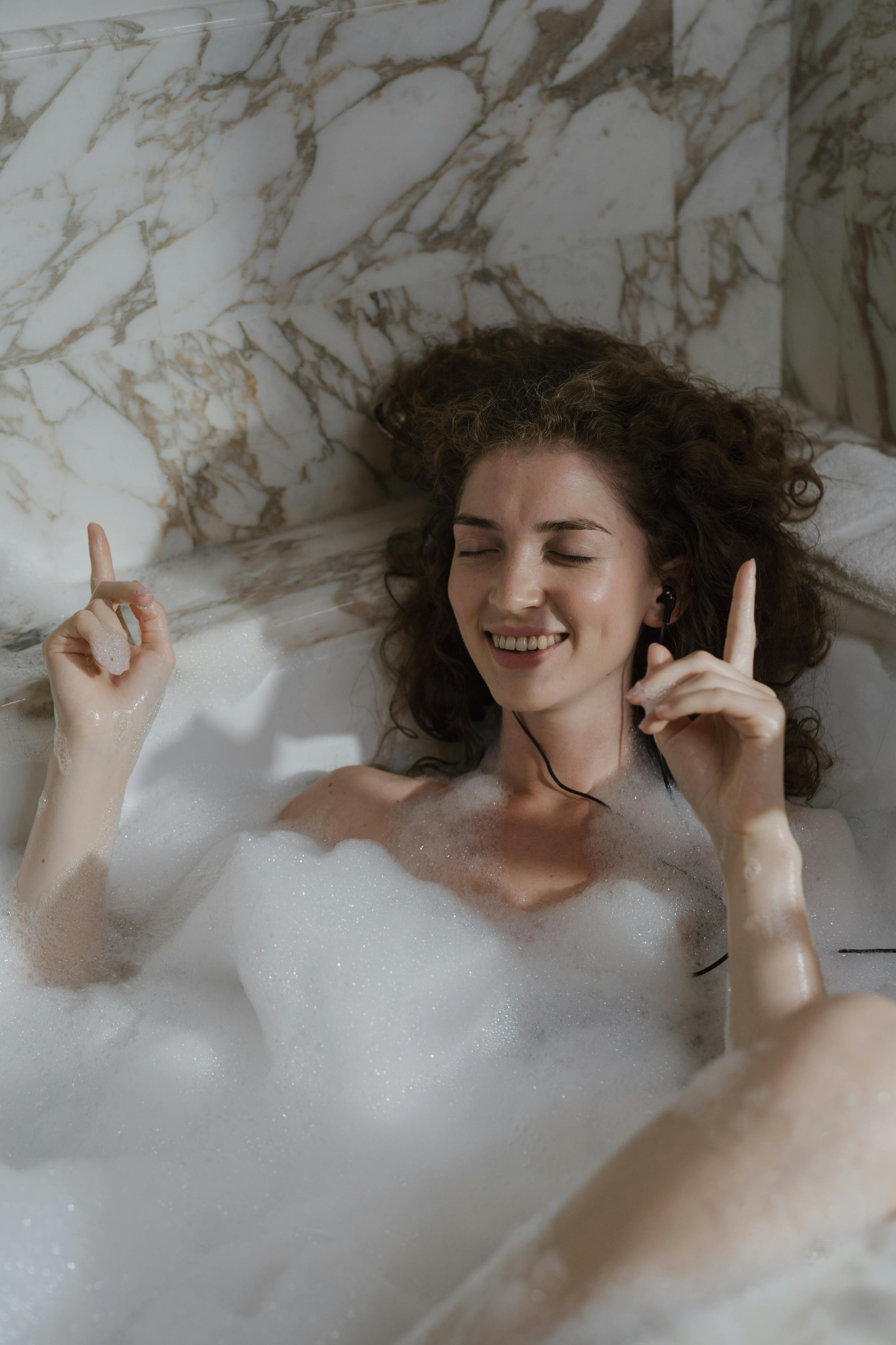 A joyful woman with curly hair indulges in music and relaxation in a luxurious bubble bath.