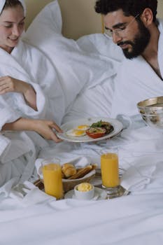 Couple enjoying a romantic breakfast in bed, wearing bathrobes, with orange juice and croissants.