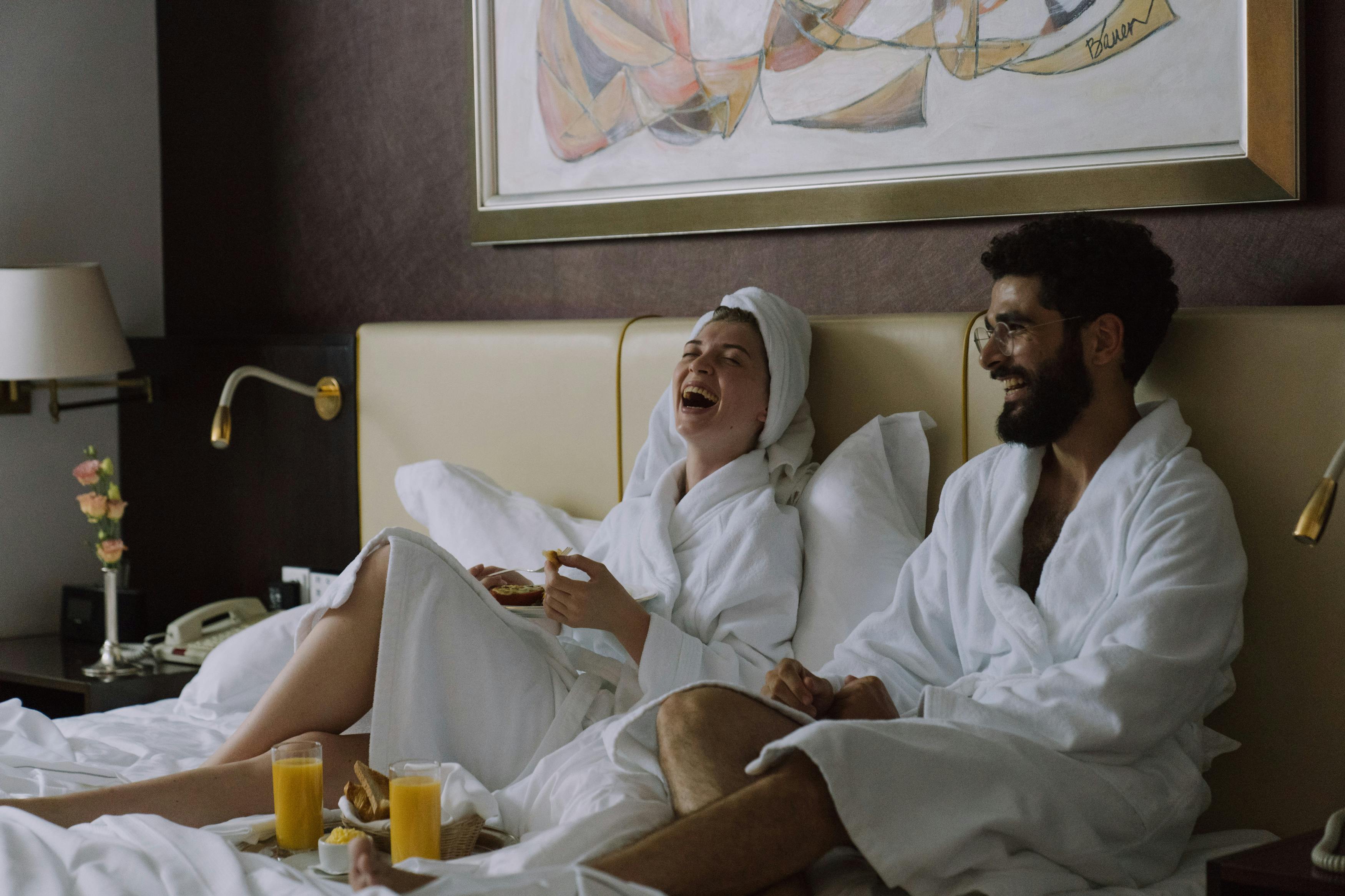 A joyful couple in bathrobes laughing while having breakfast in bed indoors.