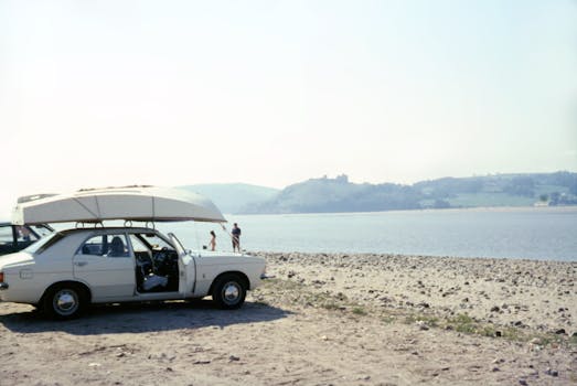 A 1970s white car with a canoe on the roof parked by a scenic beach.