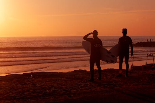 Silhouetted surfers with boards watching the sun set at Costa da Caparica, Portugal.