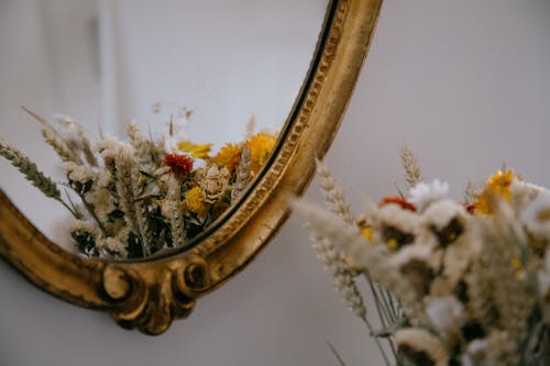 Through decorated mirror of bunch blooming flowers with dry herbs on surface near white wall