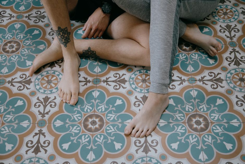 High angle of crop faceless barefoot couple sitting together with crossed tattooed legs on tiled floor