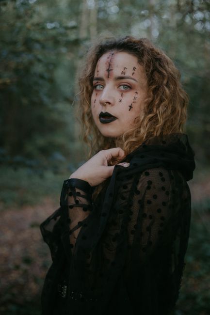 Young curly haired female with Halloween face art in black thin costume standing in green forest and looking at camera