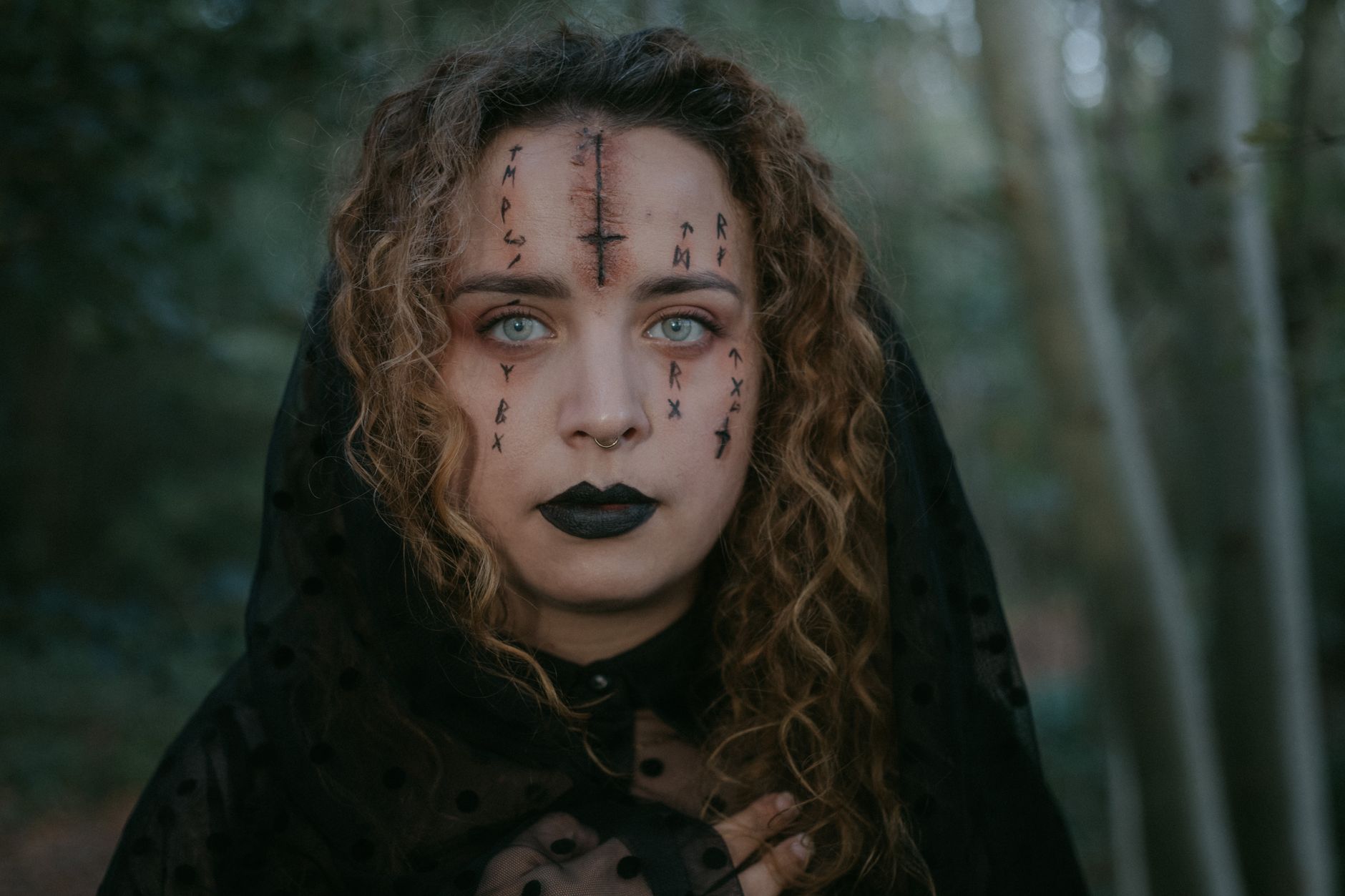 Portrait of female with spooky face art in black veil standing in forest and looking at camera