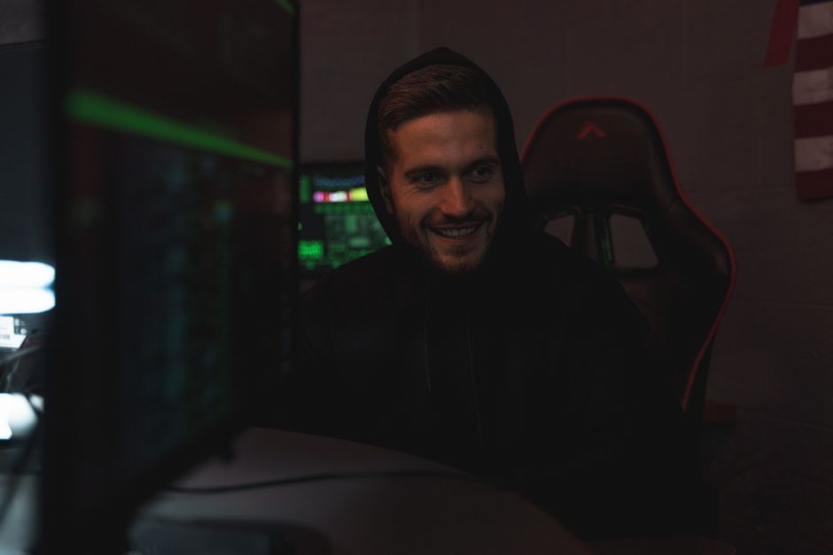 Man in a black hoodie sits in a dark room, smiling while using a computer. Tech-related setting.