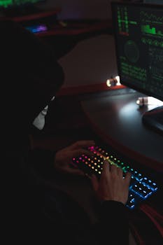 An anonymous hacker in a black hoodie typing on a colorful RGB keyboard, highlighting cybersecurity themes.