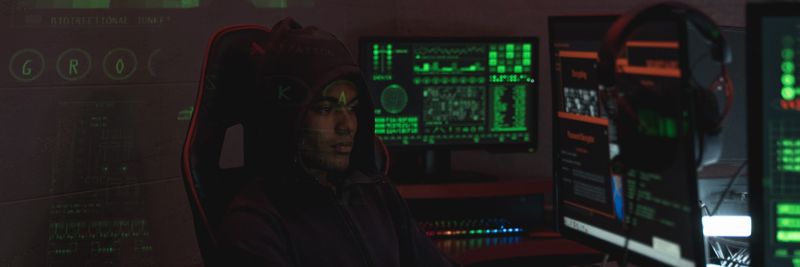 A man in a black hoodie engaged in cybersecurity work using multiple monitors indoors.
