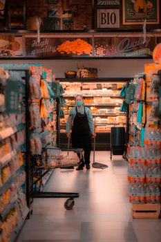 Elderly worker in mask cleans aisle of grocery store.