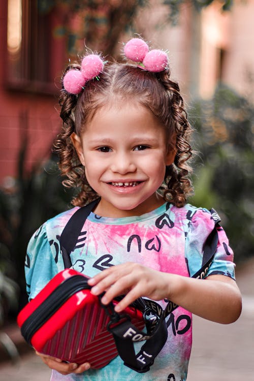Happy child standing on street and holding small red handbag while smiling and looking at camera