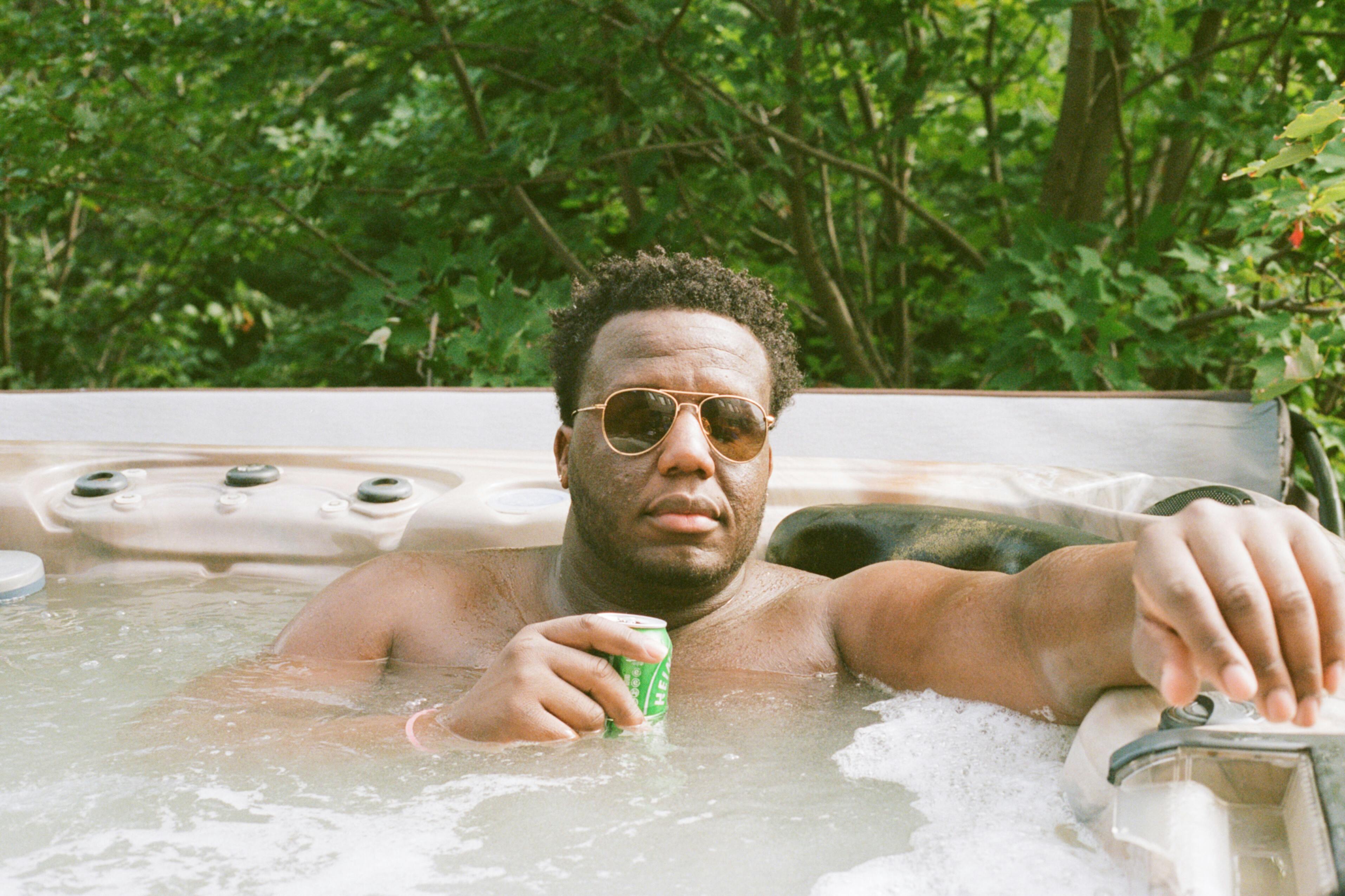 Man enjoying leisure time in hot tub outdoors, holding a drink and wearing sunglasses, surrounded by greenery.