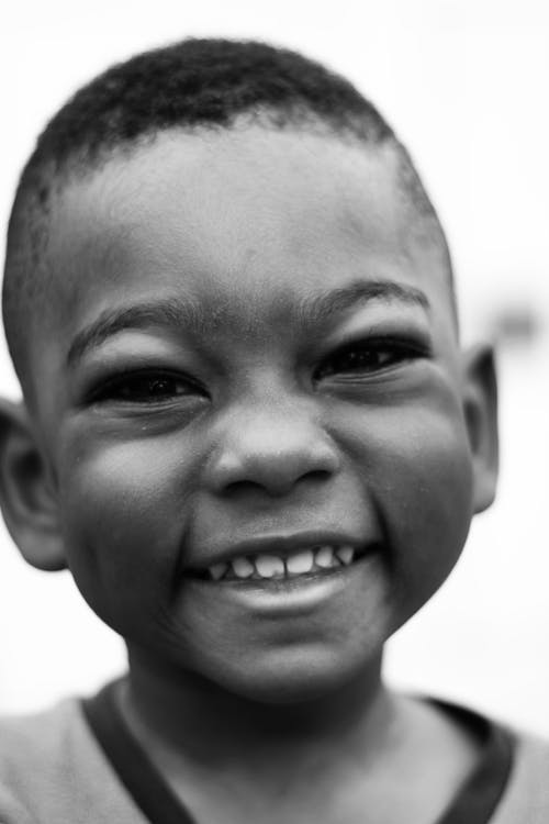 Black and white of African American little boy with short hair looking at camera with toothy smile in daylight