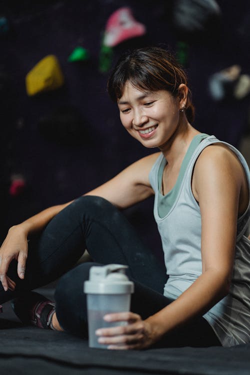 Side view of positive Asian sportswoman sitting with bottle of water and resting after bouldering workout