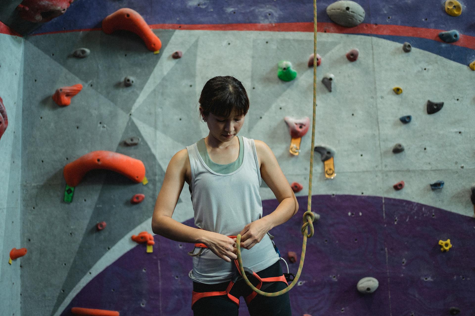 Serious Asian woman fastening safety harness and rope for climbing on wall in bouldering center