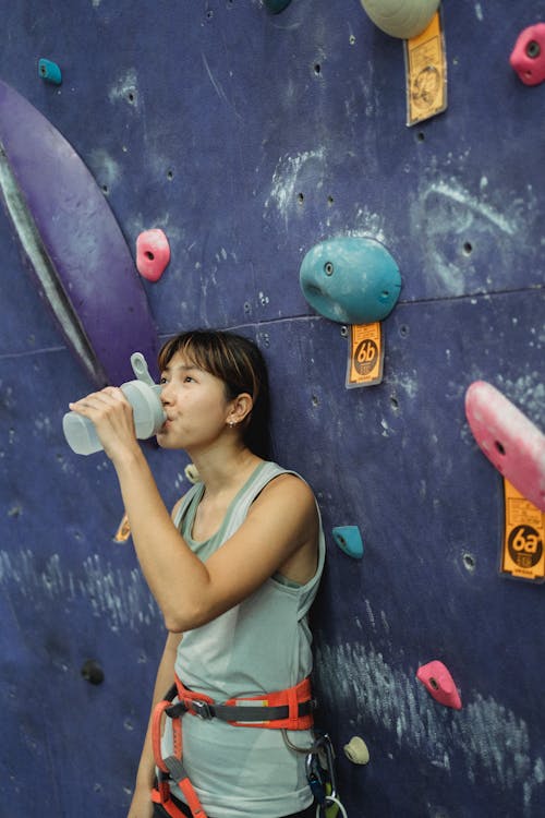 Side view young thirsty Asian female in activewear and safety harness drinking water from bottle after intense bouldering training