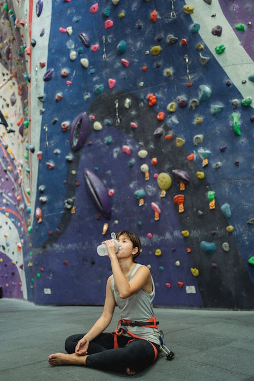 Full length tired Asian female climber in activewear sitting on floor near stiff climbing wall and drinking fresh water from bottle