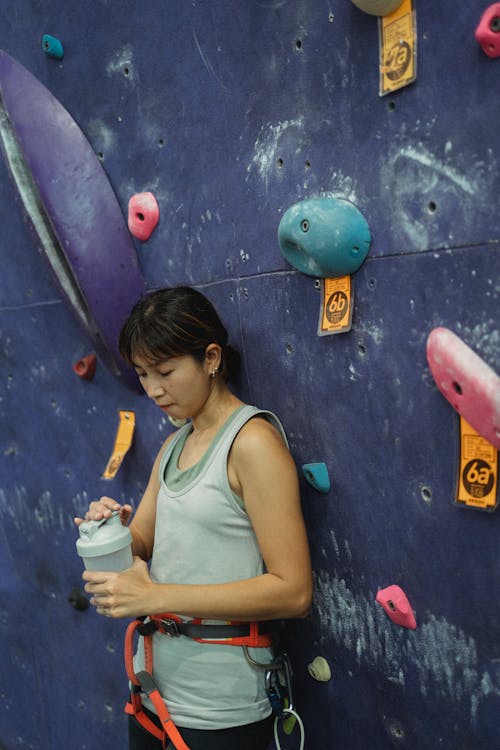 Side view fit thirsty Asian female wearing sportswear and safety harness standing near climbing wall and opening water bottle