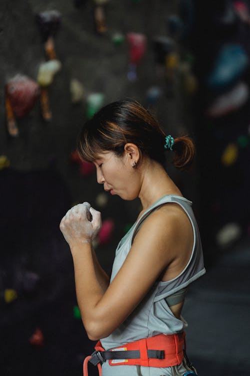 Side view of young slim ethnic female alpinist with safety belt blowing on hands in chalk before practice