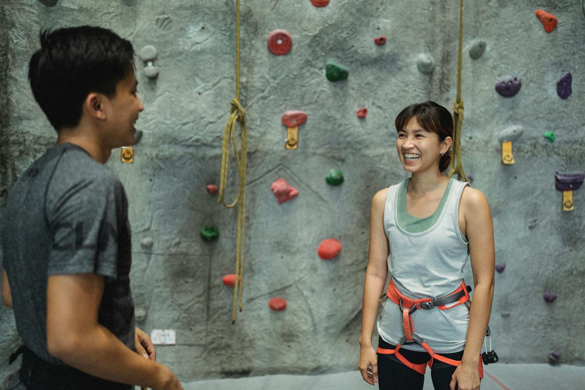Young cheerful ethnic male instructor talking to friendly female climber with safety harness while looking at each other near artificial rock