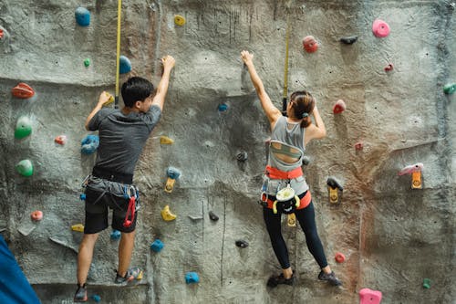 Back view of anonymous couple in activewear with safety equipment climbing rough gray rocky wall