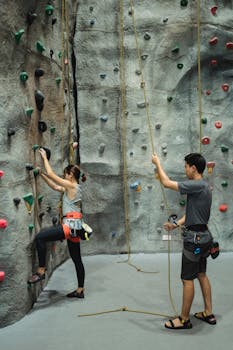 Woman rock climbing indoors with trainer belaying rope for safety.