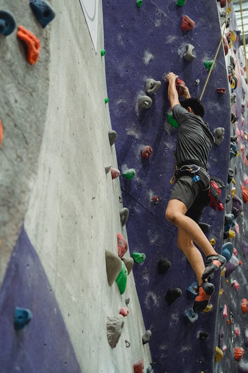 From below of anonymous male mountaineer in sportswear hanging on boulders while exercising in bouldering center