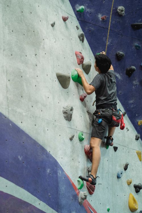 Free Back view of anonymous male climber with belay moving up on artificial wall during training in bouldering center Stock Photo