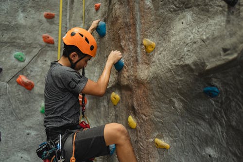 Free Side view of crop young Asian man in orange helmet and belay ascending on climbing wall during training in bouldering center Stock Photo