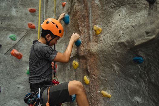 Side view of crop young Asian man in orange helmet and belay ascending on climbing wall during training in bouldering center