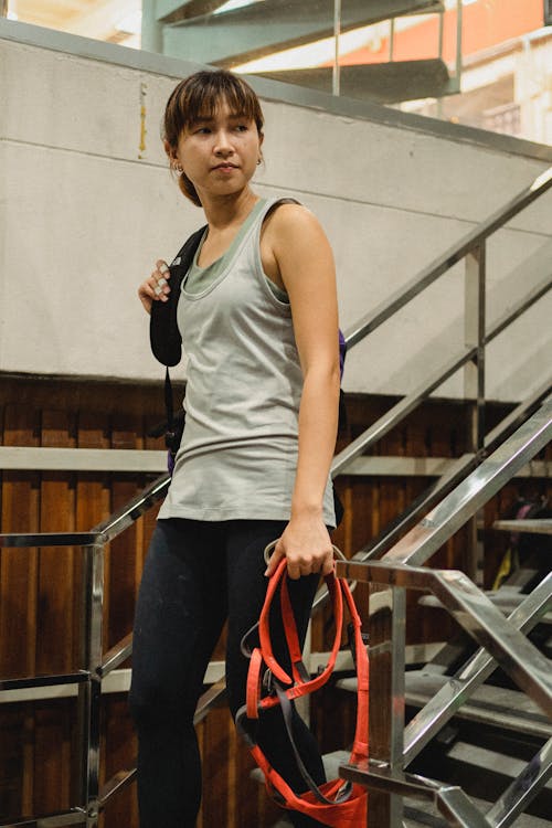 Young Asian sportswoman walking down staircase with backpack and orange safety equipment before climbing training in bouldering gym