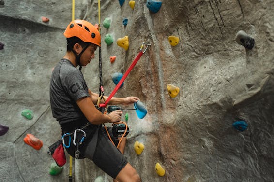 From below of side view of Asian male specialist in helmet drilling hole in blue climbing hold while hanging on safety rope near bouldering wall
