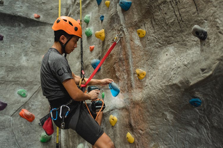 Asian Technician Making Hole In Climbing Hold With Screwdriver While Hanging On Safety Rope