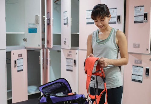 Smiling young Asian woman in activewear standing in locker room and preparing safety equipment for training in climber hall