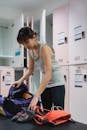 Young sportive Asian lady getting dark sneakers out from gym bag while standing near bench with safety belt  in changing room before training