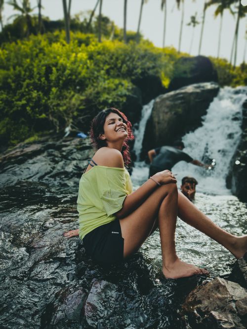 Free A woman enjoys the summer by sitting on rocks near a cascading waterfall, showcasing happiness and relaxation. Stock Photo