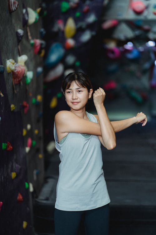 Smiling Asian woman in activewear warming up hands before practicing climbing on artificial wall in bouldering gym