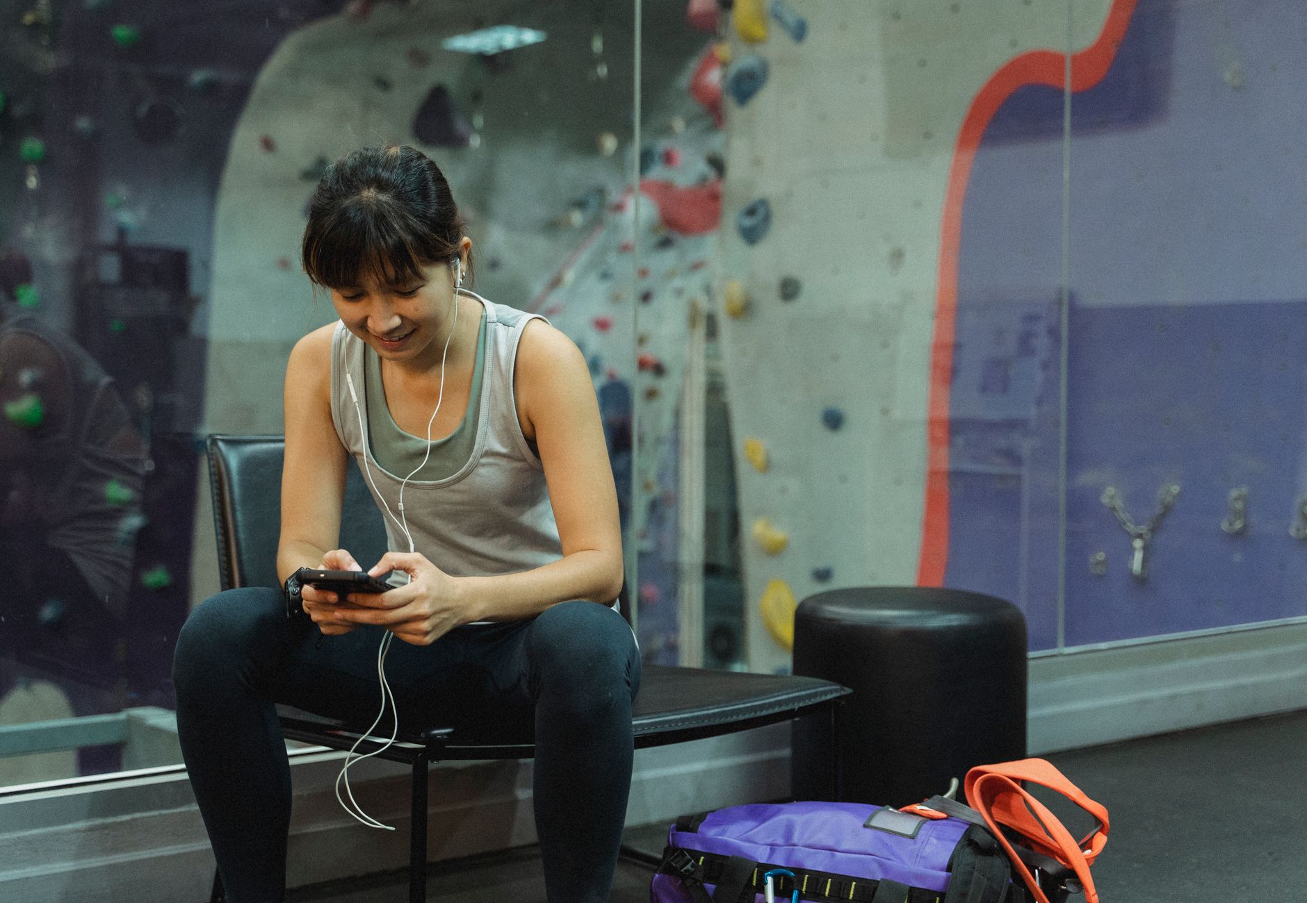 Happy young sporty ethnic lady in activewear sitting on bench and messaging on mobile phone while listening to music in earphones during break in gym