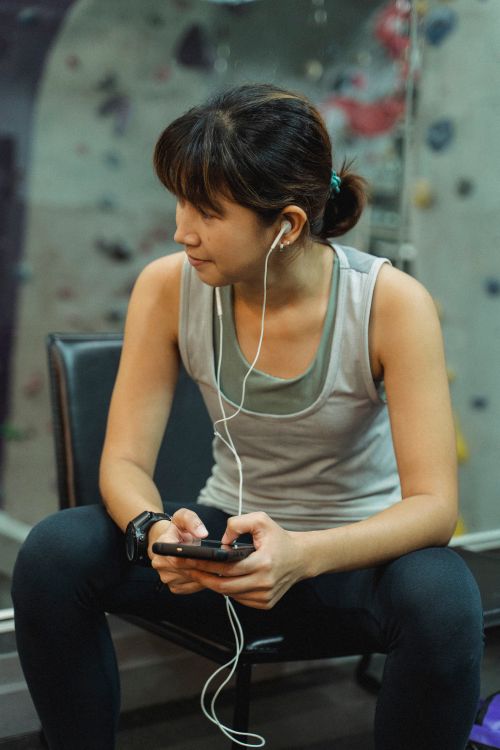 Free Young sporty Asian lady in activewear and earphones browsing mobile phone while relaxing on bench after workout in climbing gym Stock Photo