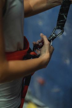 Side view of crop faceless female alpinist fastening safety harness and belay rope before climbing wall in gym