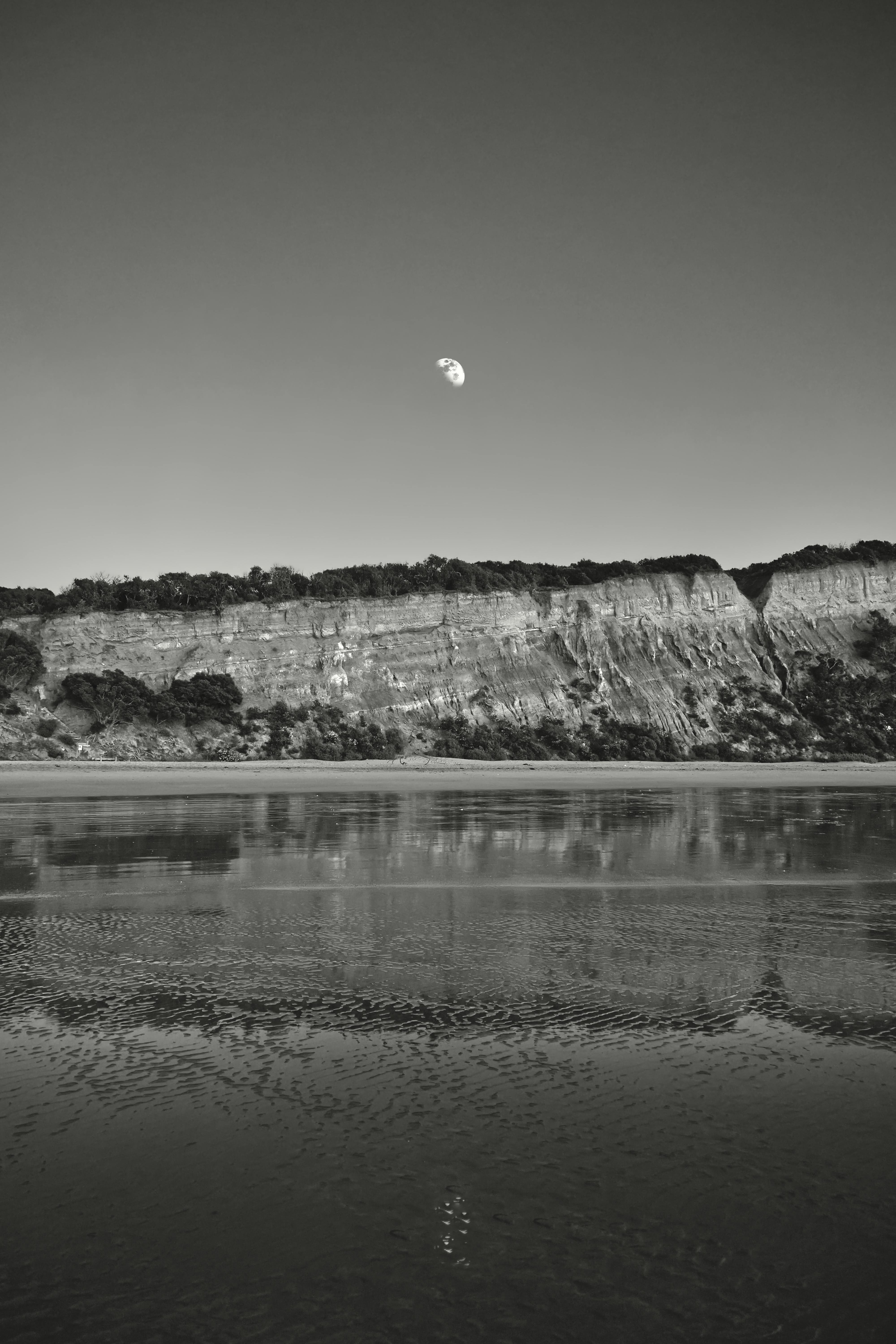 Black and white image of Bells Beach cliffs with moon and water reflection.