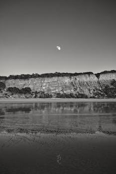 Black and white image of Bells Beach cliffs with moon and water reflection.
