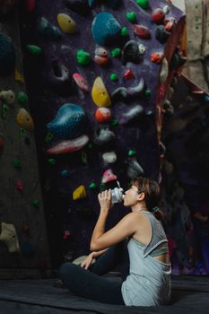 Full body side view of Asian female mountaineer drinking water while sitting with crossed legs at artificial climbing wall during break