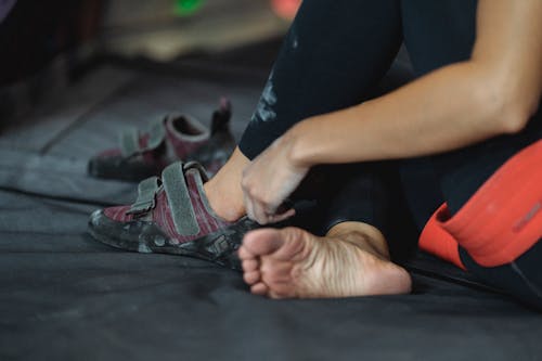 Free Side view of unrecognizable barefoot mountaineer sitting on mat with crossed legs while putting on rock climbing shoes during training in gym on blurred background Stock Photo