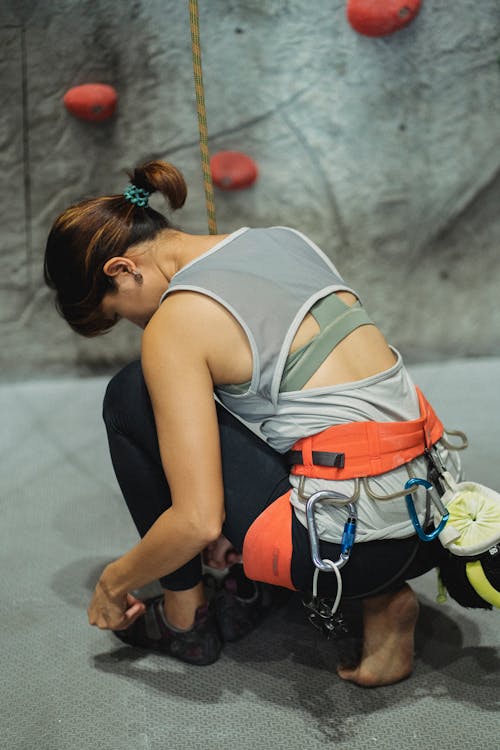 Free Full body back view of anonymous female mountaineer with belay squatting while putting on rock climbing shoes near artificial wall with grips Stock Photo