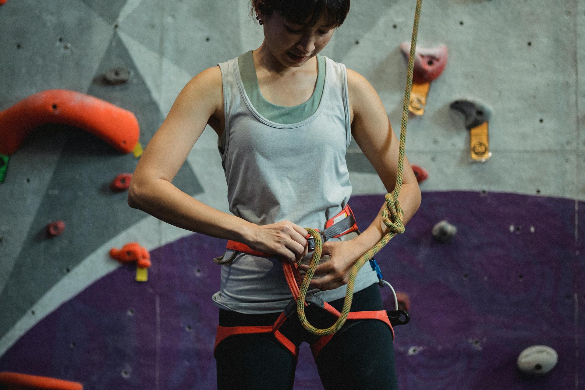 Crop young Asian sportswoman in activewear and safety sit harness preparing belay for climbing training in gym