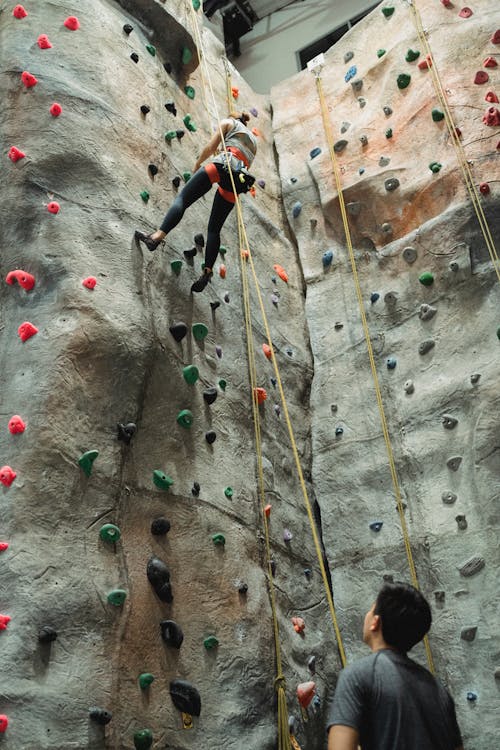 From below of unrecognizable male instructor looking at female climber practicing bouldering on colorful artificial rock in climbing gym