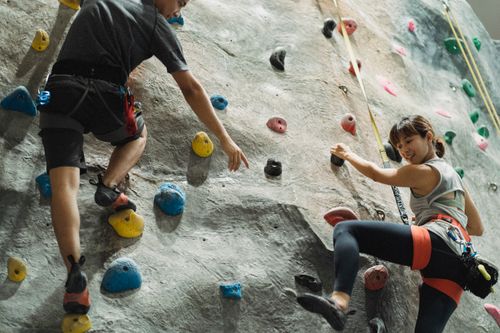 Free Back view of crop anonymous male trainer helping young Asian female athlete hanging on belay during climbing training in gym Stock Photo